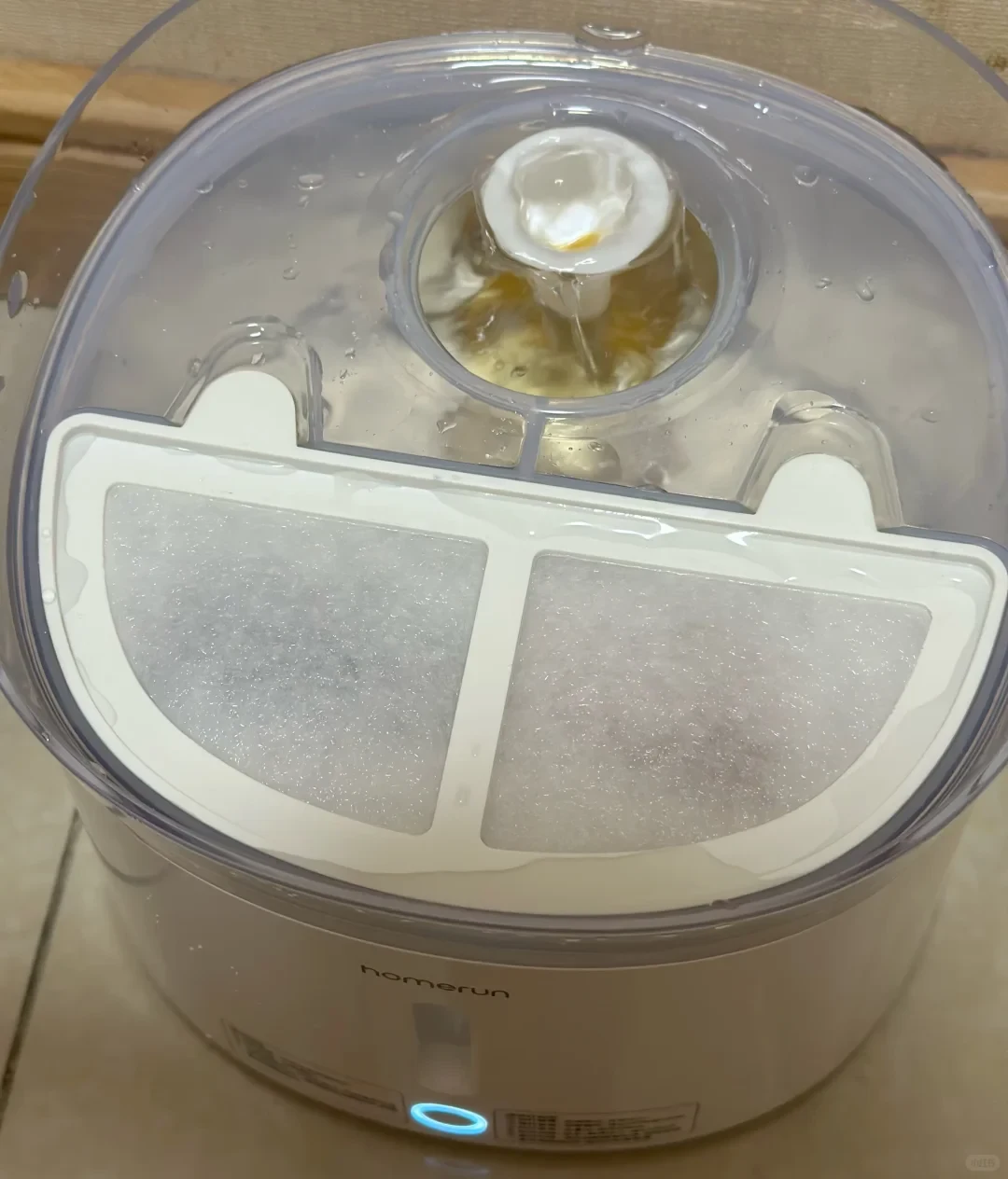 Top view of an operational Homrun pet water fountain with bubbling water, a visible filter compartment, clear plastic top, and an illuminated blue indicator light on the white base.