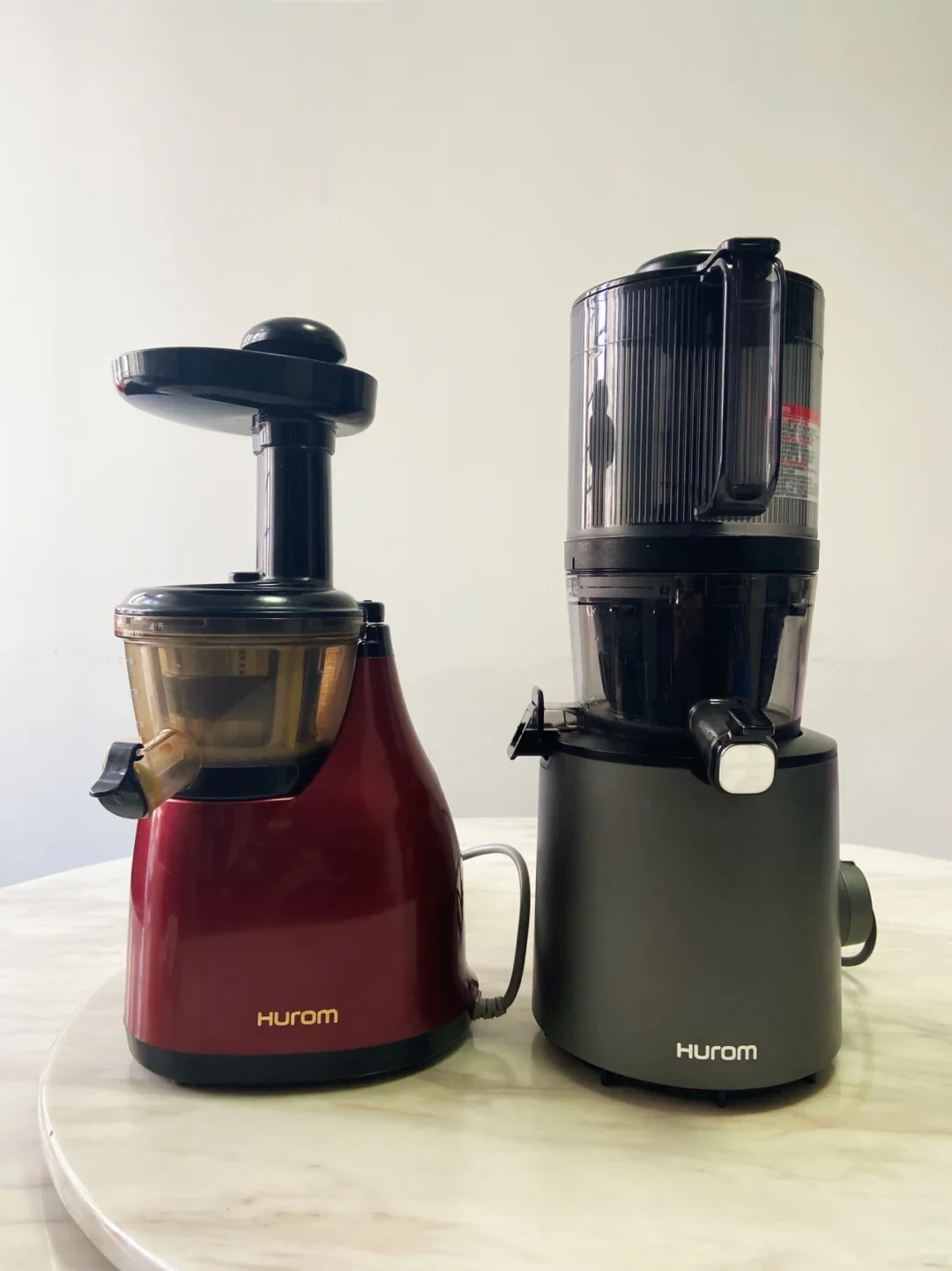 Two Hurom juicers stand side by side on a light-colored table: a burgundy model with an amber transparent bowl on the left and a dark gray model with a tall ribbed hopper on the right.