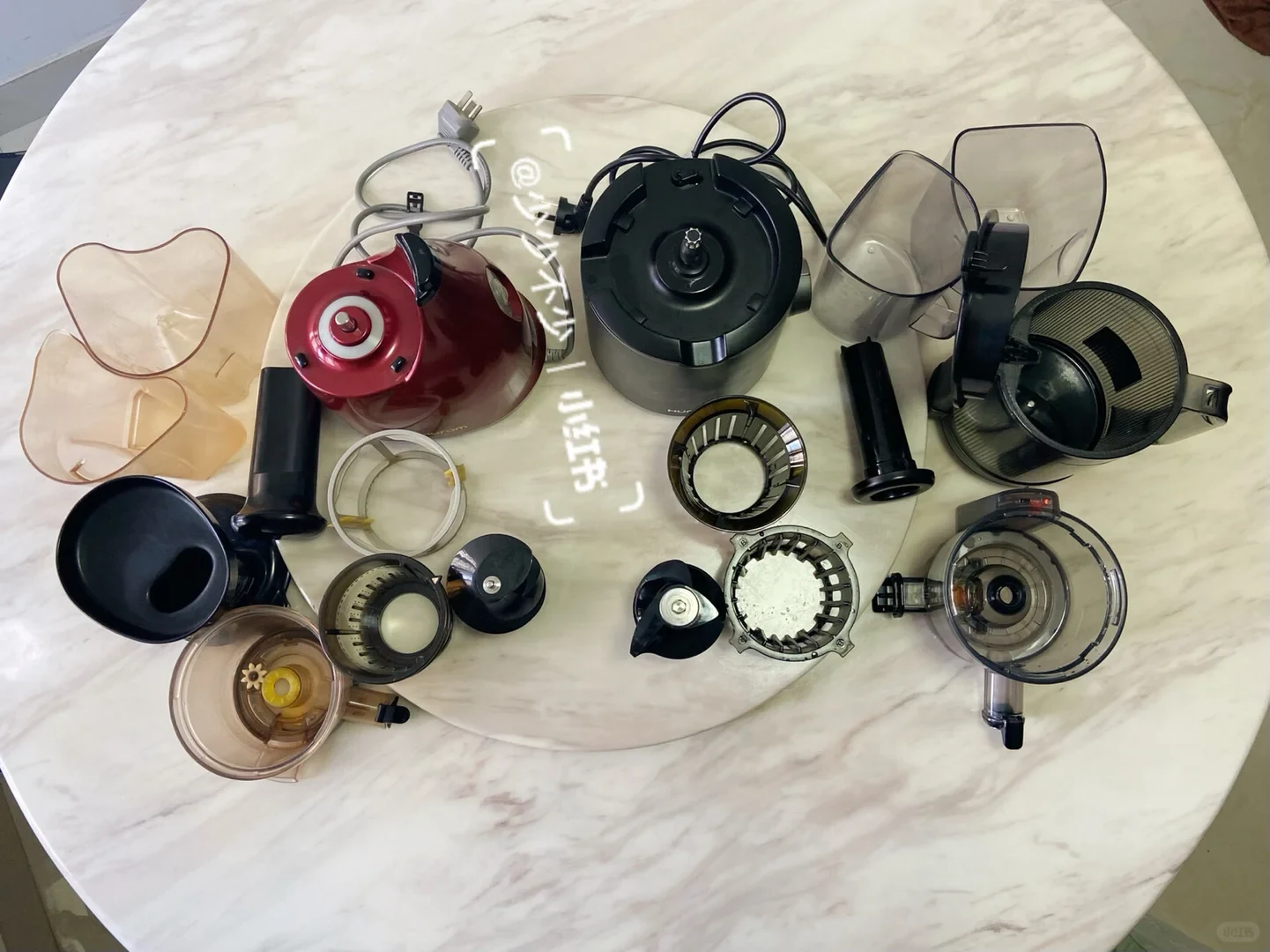 A red-and-black juicer is shown disassembled on a round marble table, with its motor base, power cord, clear containers, filters, lids, and other removable parts laid out around it.