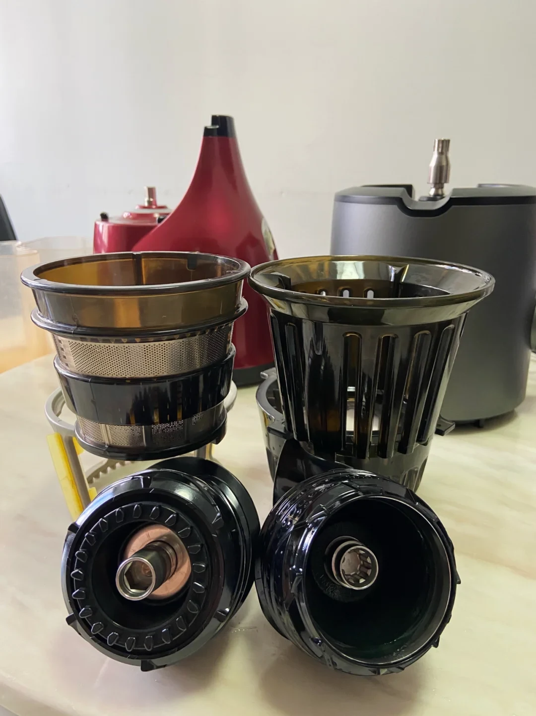 Disassembled juicer parts are arranged on a countertop, including two black filter or strainer baskets and two round black components, with the juicer’s red and gray motor housings behind them.