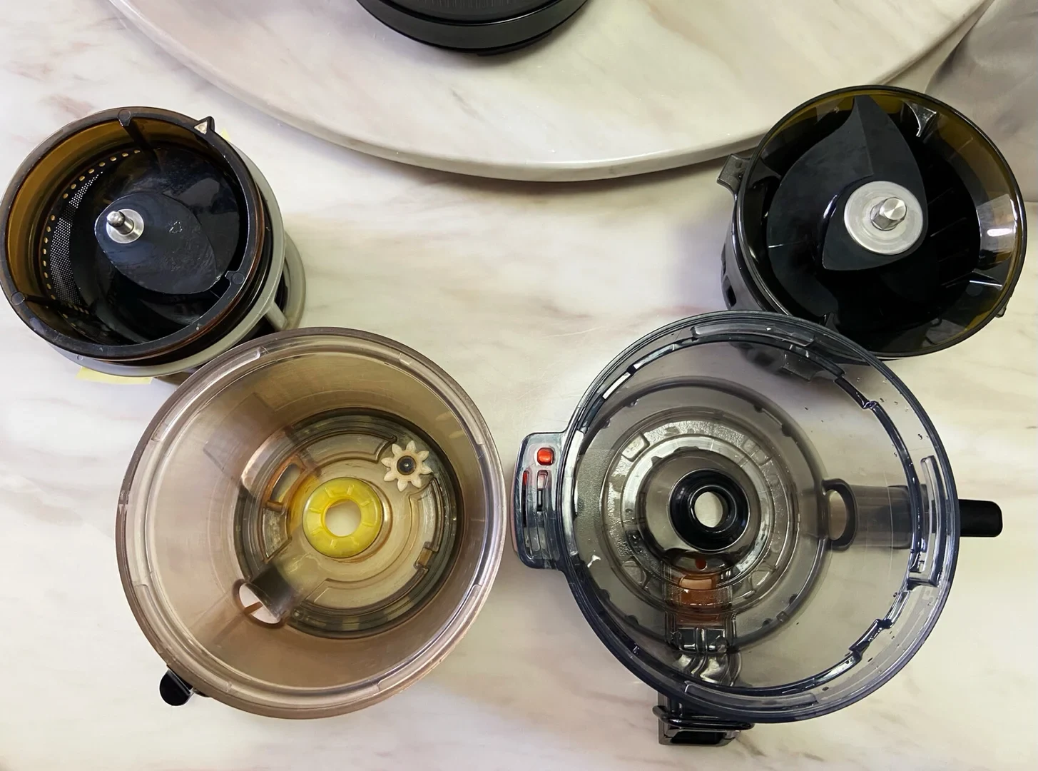 Four disassembled juicer components are arranged on a light marble surface, including two transparent collection bowls and two black circular parts with central metal fittings.