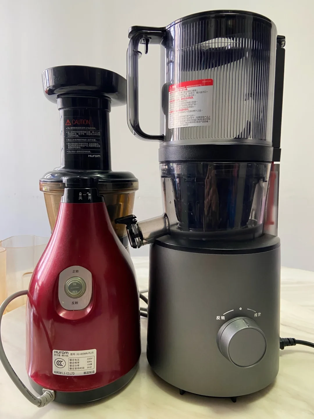 A red Hurom slow juicer and a taller gray juicer stand side by side on a countertop, with Chinese-labeled controls and instruction stickers visible.