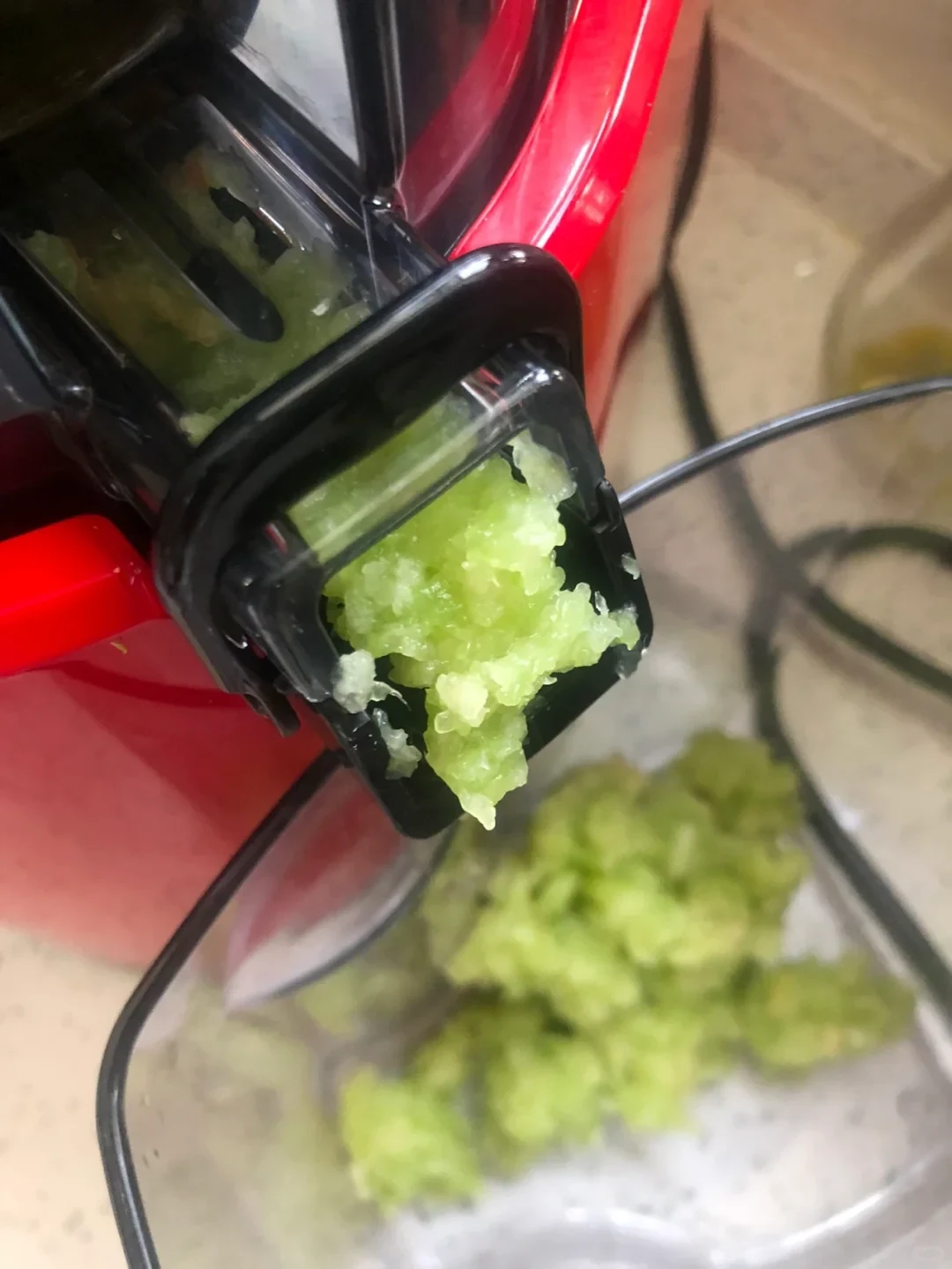 A red-and-black juicer presses pale green produce pulp through its outlet into a clear bowl containing more pulp.
