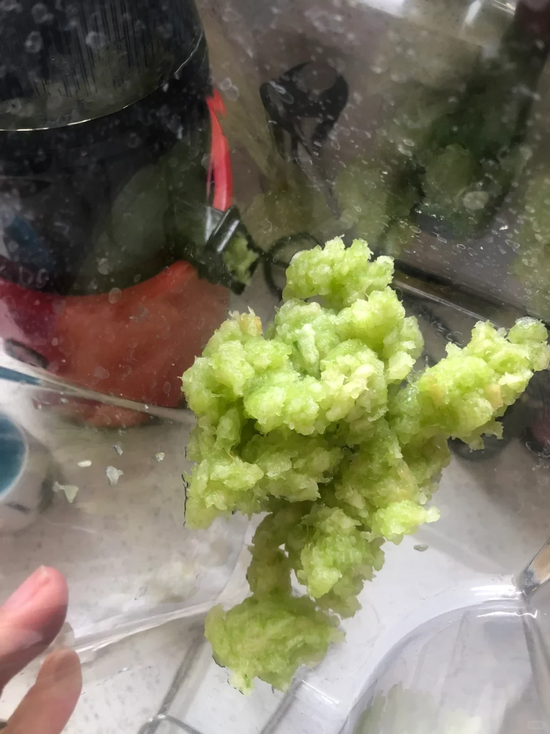 A close-up of a kitchen appliance dispensing a clump of moist, shredded light-green food into a clear container, with a hand holding the container at the lower left.