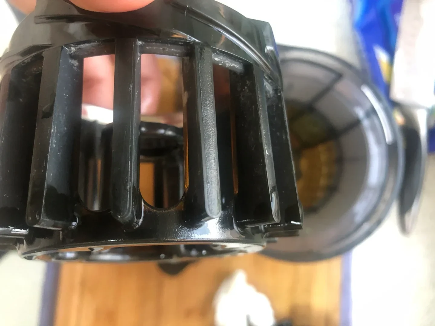 Close-up of a hand holding a black plastic cylindrical component with several vertical slotted openings and a curved rim; another round black component is blurred in the background.