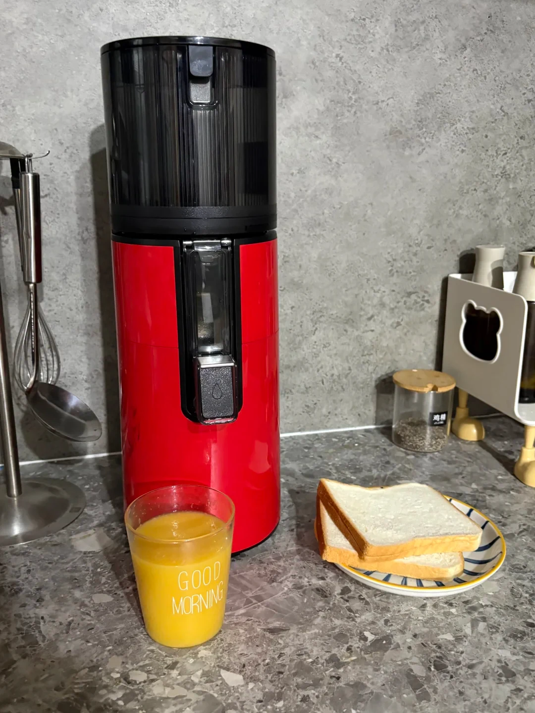 A red-and-black countertop juicer stands on a kitchen counter beside a glass of orange juice and a plate of sliced white bread. A whisk and ladle hang at left, and small containers sit at right.