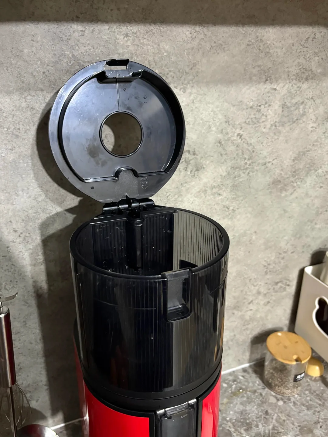 A red-and-black countertop appliance with its round lid open, revealing an empty, vertically ribbed black container and a circular opening in the lid. It stands on a stone countertop.