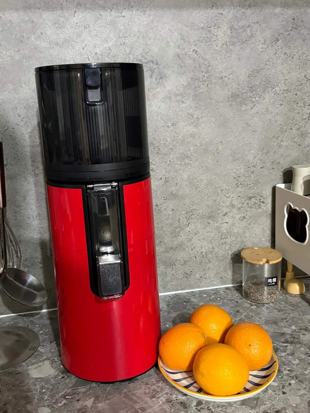 A tall red-and-black countertop appliance with a clear front window stands beside a plate of oranges on a kitchen counter.