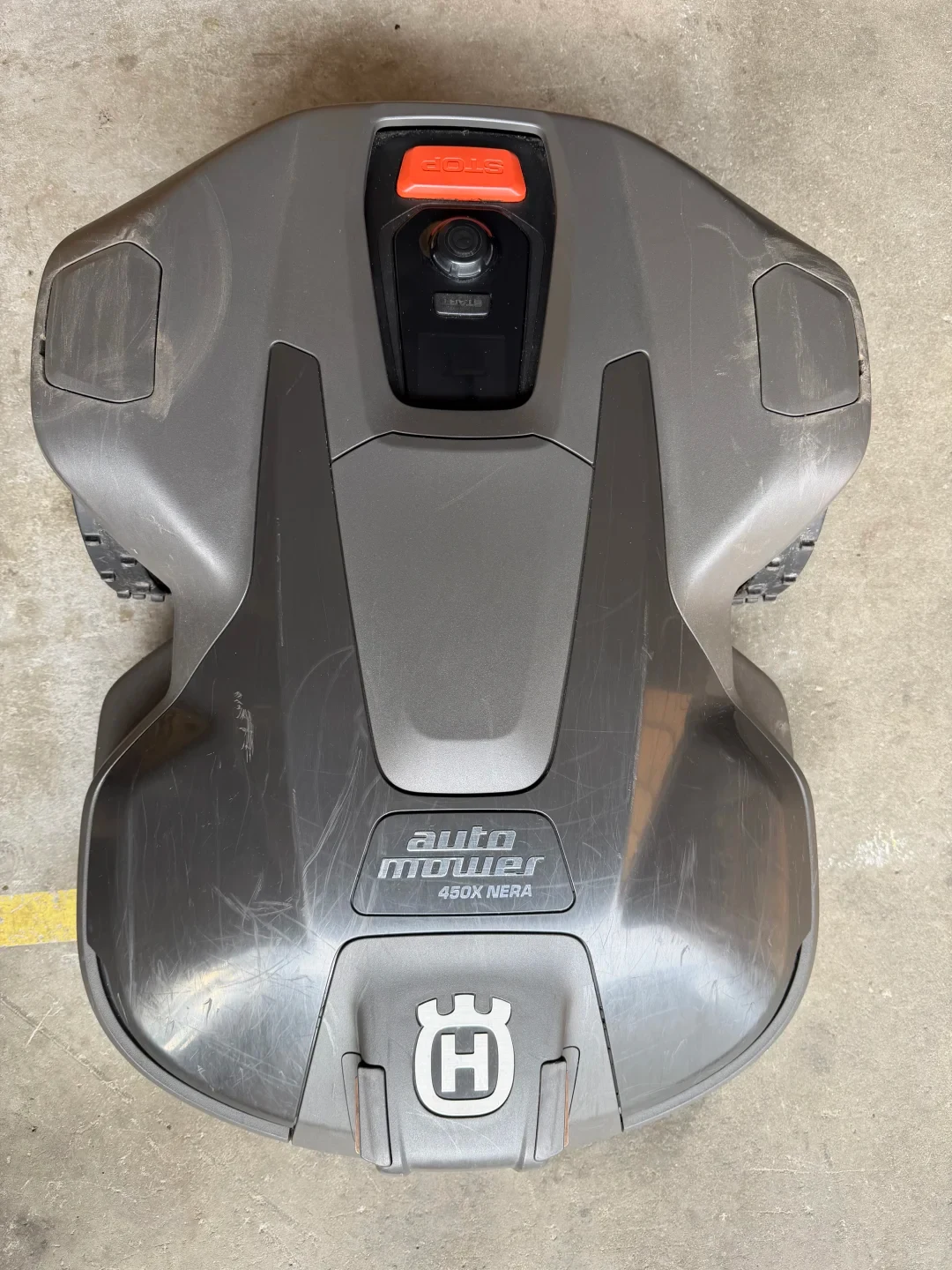 Top-down view of a grey Husqvarna Automower 450X NERA robotic lawn mower on a concrete floor, showing surface scuffs, an orange emergency stop button, control console, and the brand emblem.