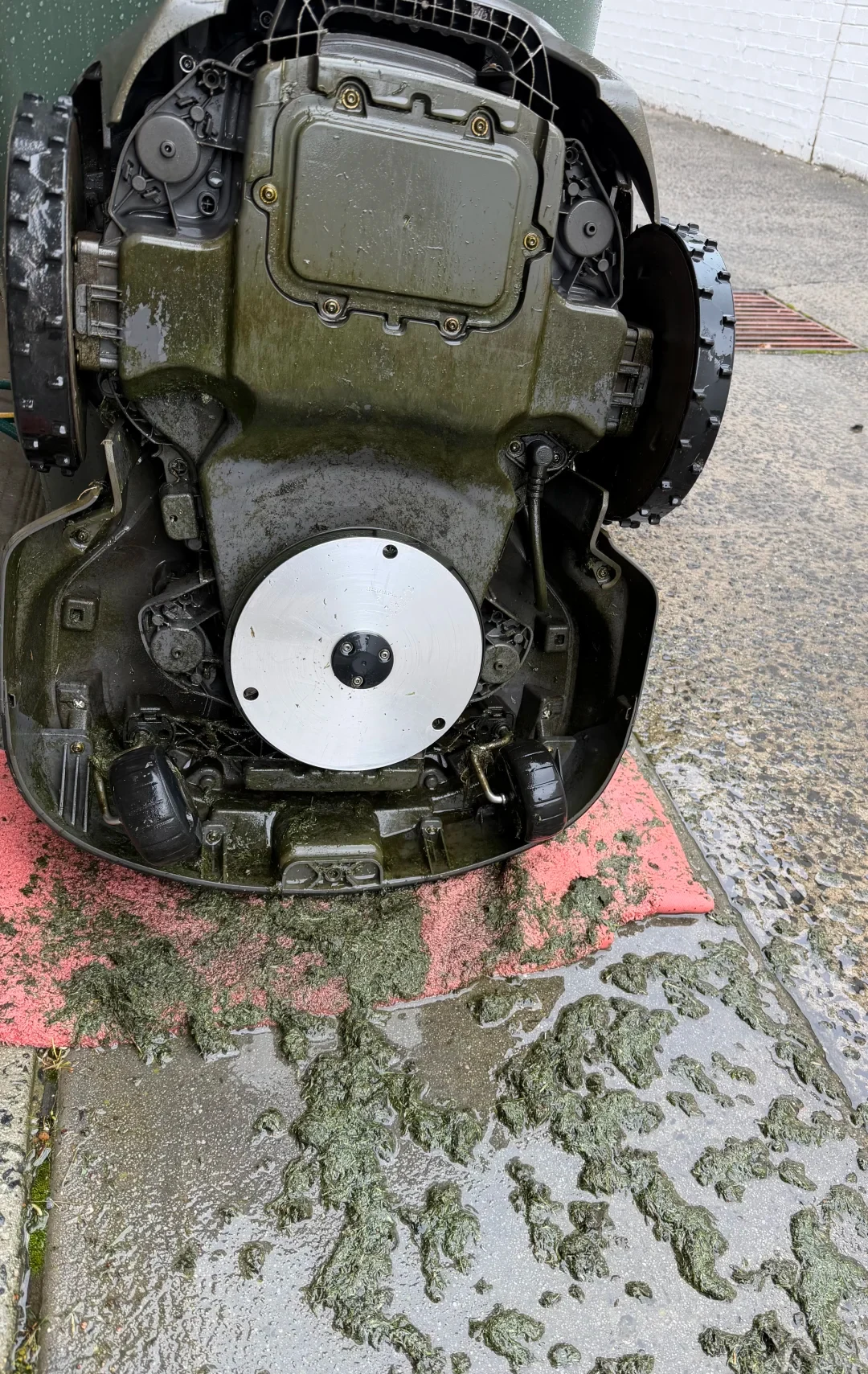 Underside view of an overturned robotic lawn mower sitting on a wet concrete surface next to a pink towel covered in wet grass clippings, showing its wheels, chassis, and circular silver cutting disc.