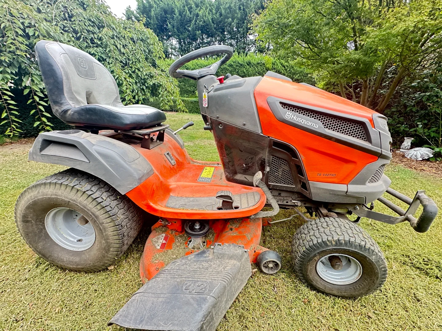 A side view of an orange and grey Husqvarna TS 142TXD riding lawn mower parked on a grass lawn with dense green trees and hedges in the background.