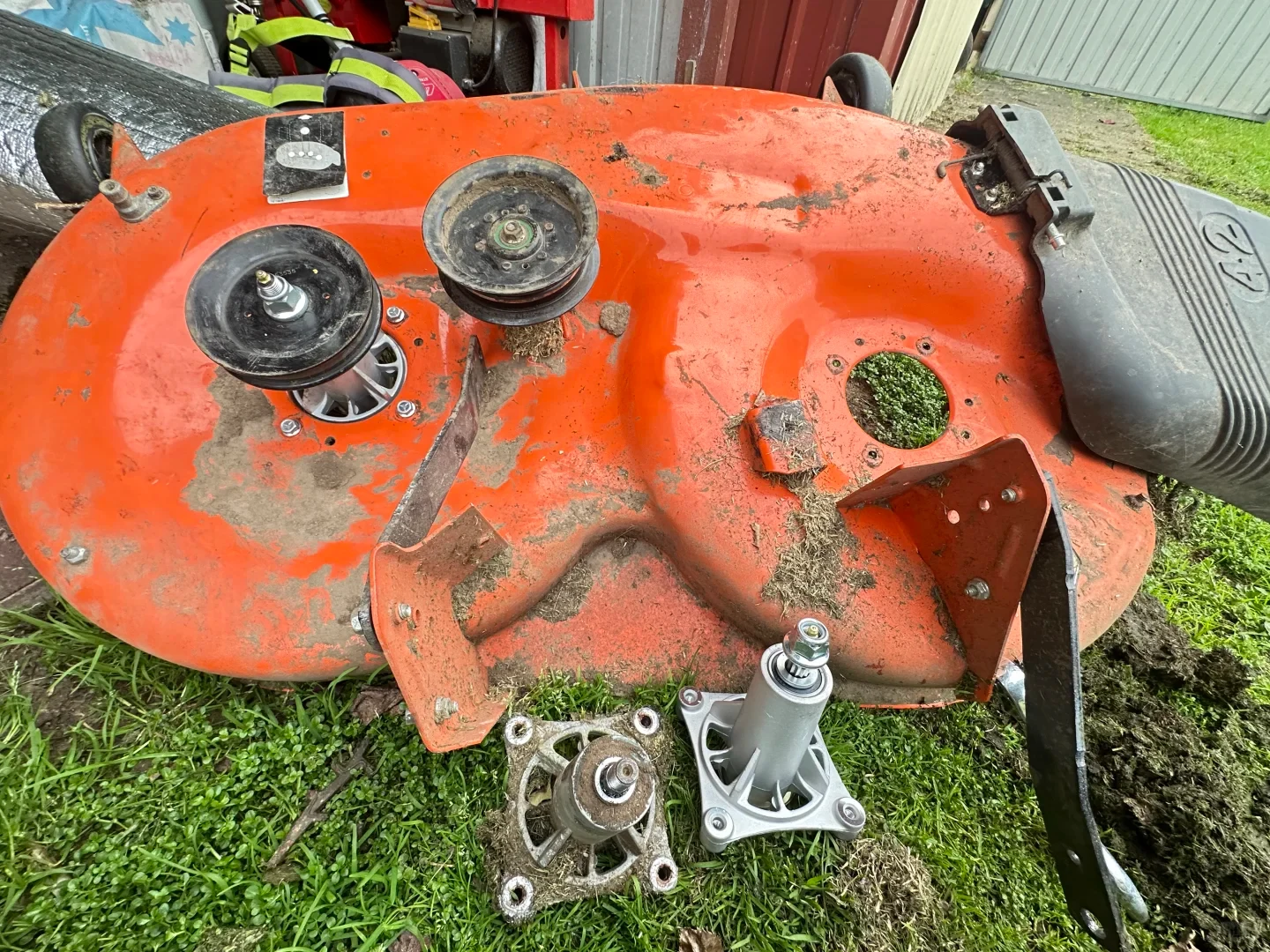 An orange riding mower cutting deck resting on a grassy lawn during repair, showing an installed spindle and pulley on the left and an empty mounting hole on the right. In the foreground below the deck lie an old, worn spindle assembly covered in dirt and a brand-new silver replacement spindle.
