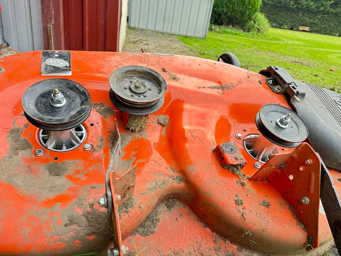 Top view of an orange riding mower cutting deck resting on the ground, featuring black spindle pulleys, an idler pulley clogged with dry grass, and a belt diagram sticker.