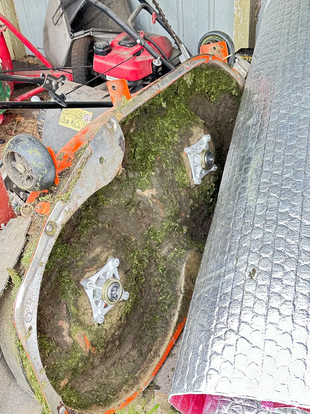 Underside of an orange riding lawn mower cutting deck tilted upright, covered in accumulated dried grass clippings and soil, showing two newly installed silver blade spindle assemblies with the cutting blades removed.