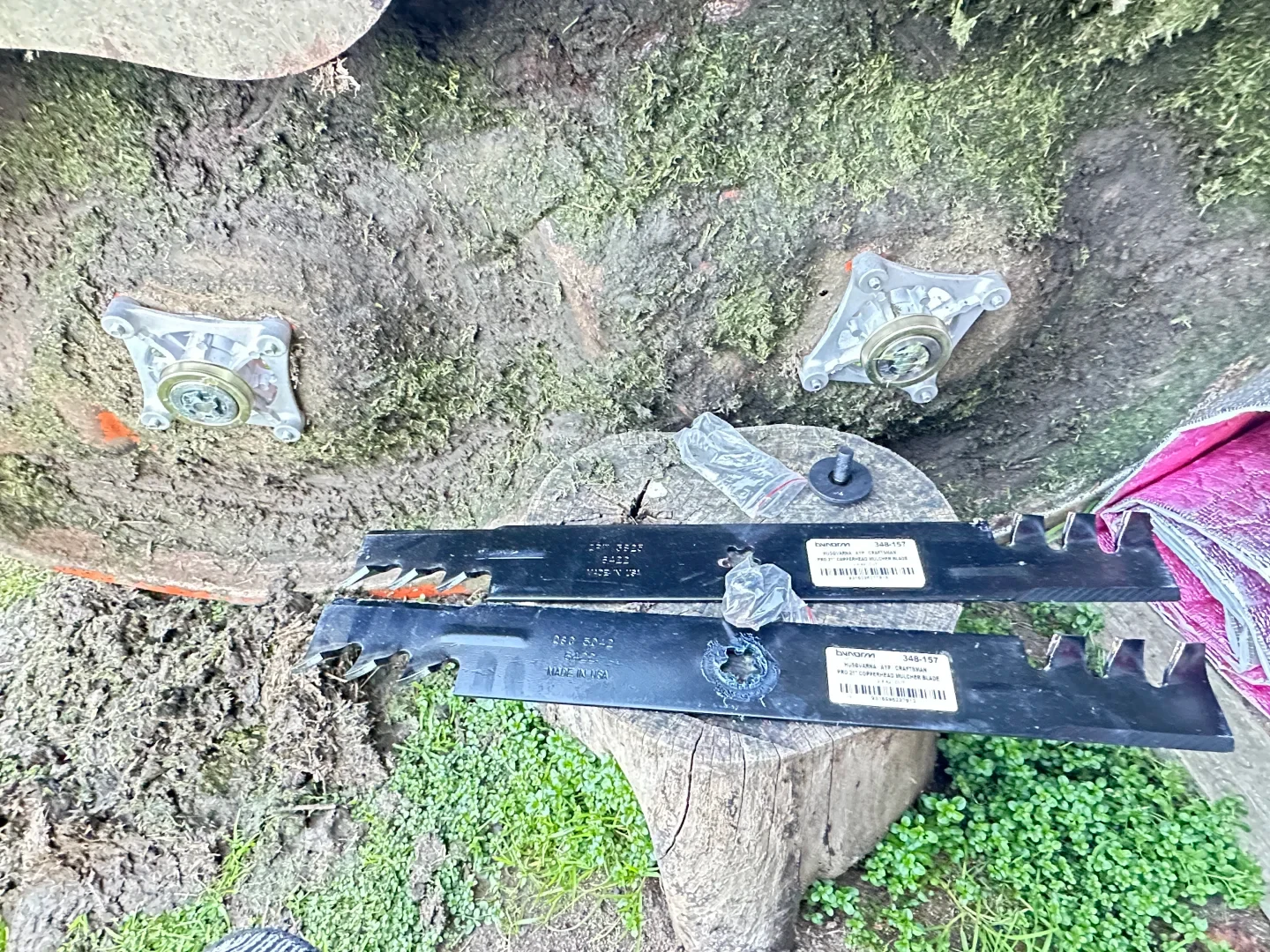 Underside of a moss- and grass-caked lawnmower deck showing two metal spindle assemblies installed, with two new black serrated mulching blades and mounting bolts resting on a tree stump below.