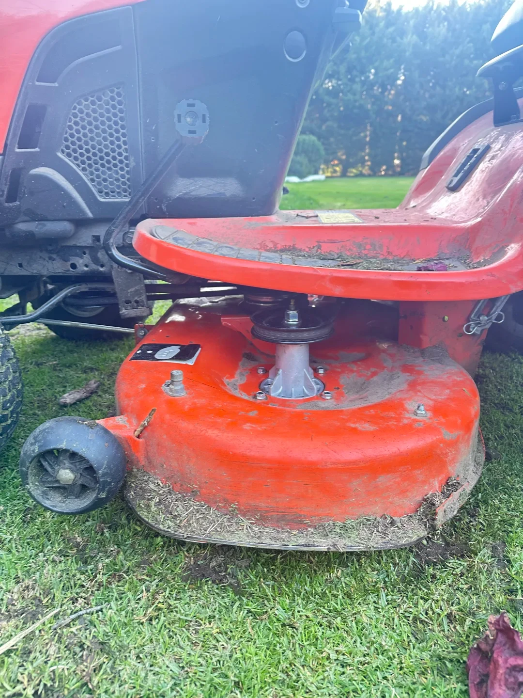 A close-up view of the red cutting deck on an orange riding lawn mower sitting on grass, showcasing a newly installed silver spindle assembly with a pulley and an anti-scalp deck wheel.