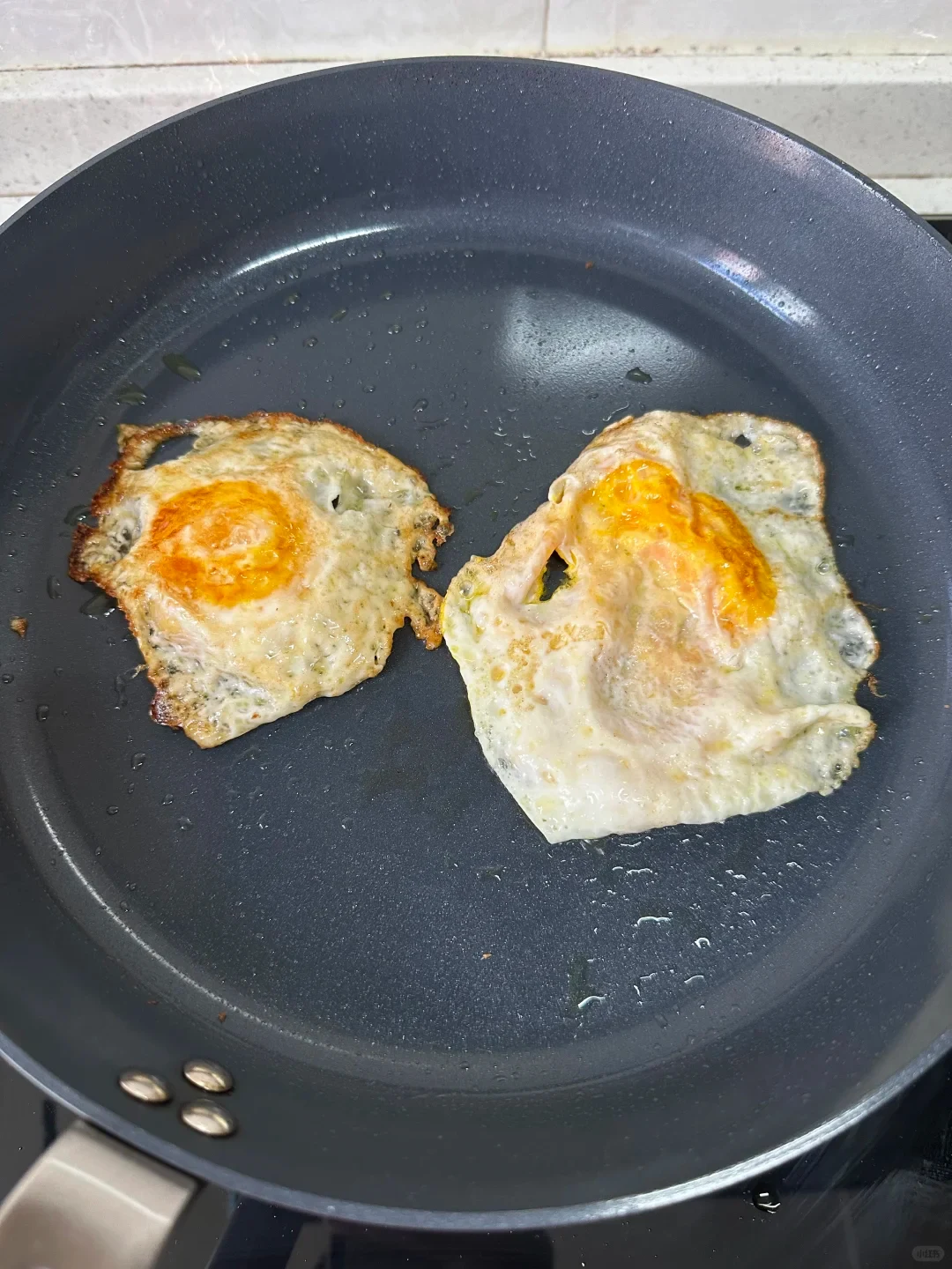 Two fried eggs cooking inside a large dark nonstick frying pan.
