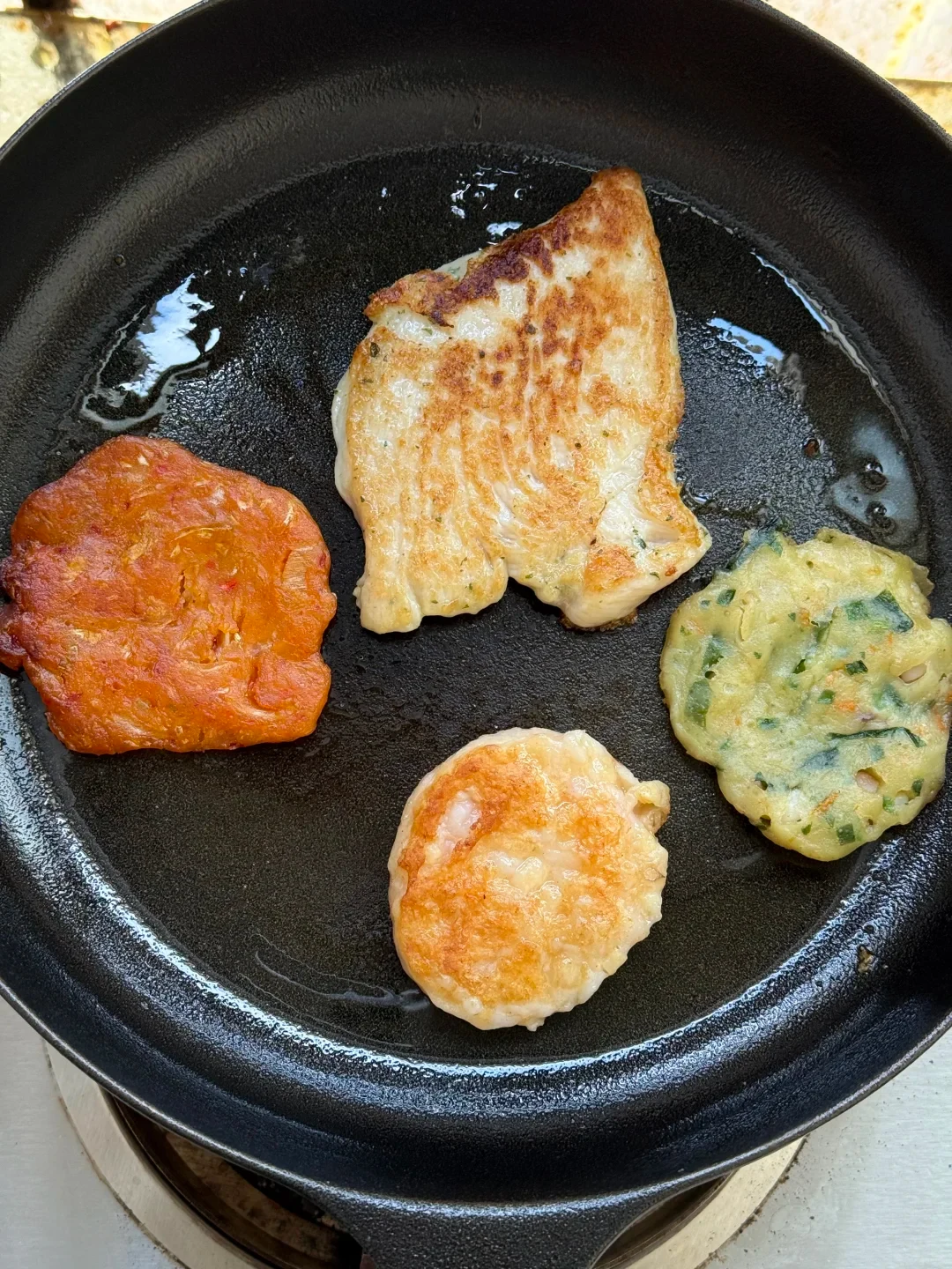 A browned fillet and several small pancakes cooking in oil in a black skillet.