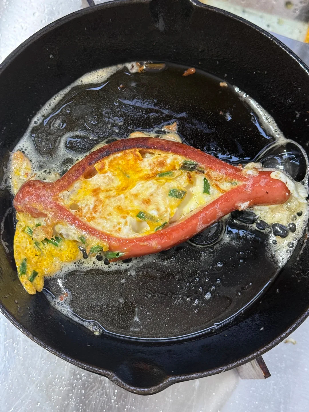 A hot dog split down the middle into an oval shape and filled with cooking egg and green onions, frying in a black cast-iron skillet.