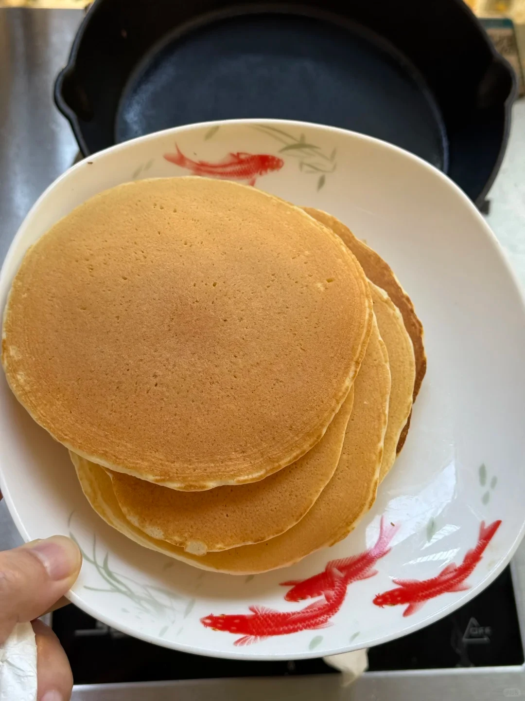A stack of evenly golden-brown round pancakes on a white ceramic plate painted with red goldfish designs, held by a hand in a kitchen setting.