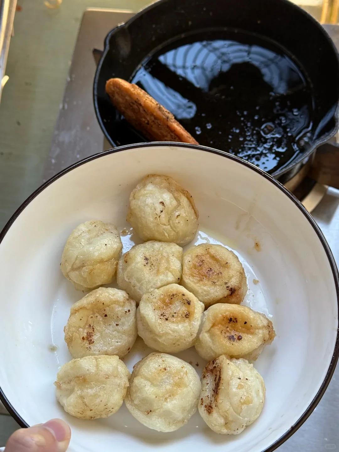 A white bowl containing small, round fried pastries or dumplings, with a cast-iron skillet filled with cooking oil in the background.