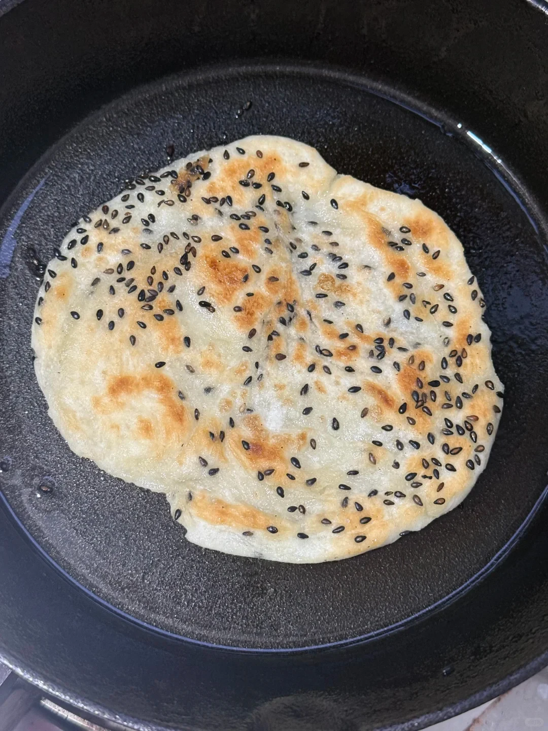 A golden-brown flatbread topped with black sesame seeds frying in a round black cast-iron pan.
