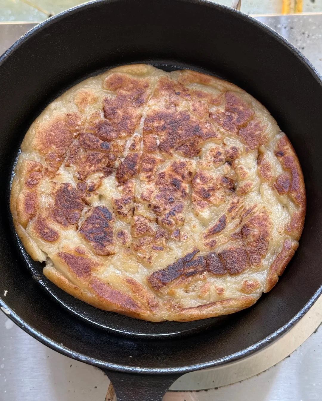 A thick round pancake with a golden-brown, cracked crispy crust frying in oil inside a black cast-iron skillet.