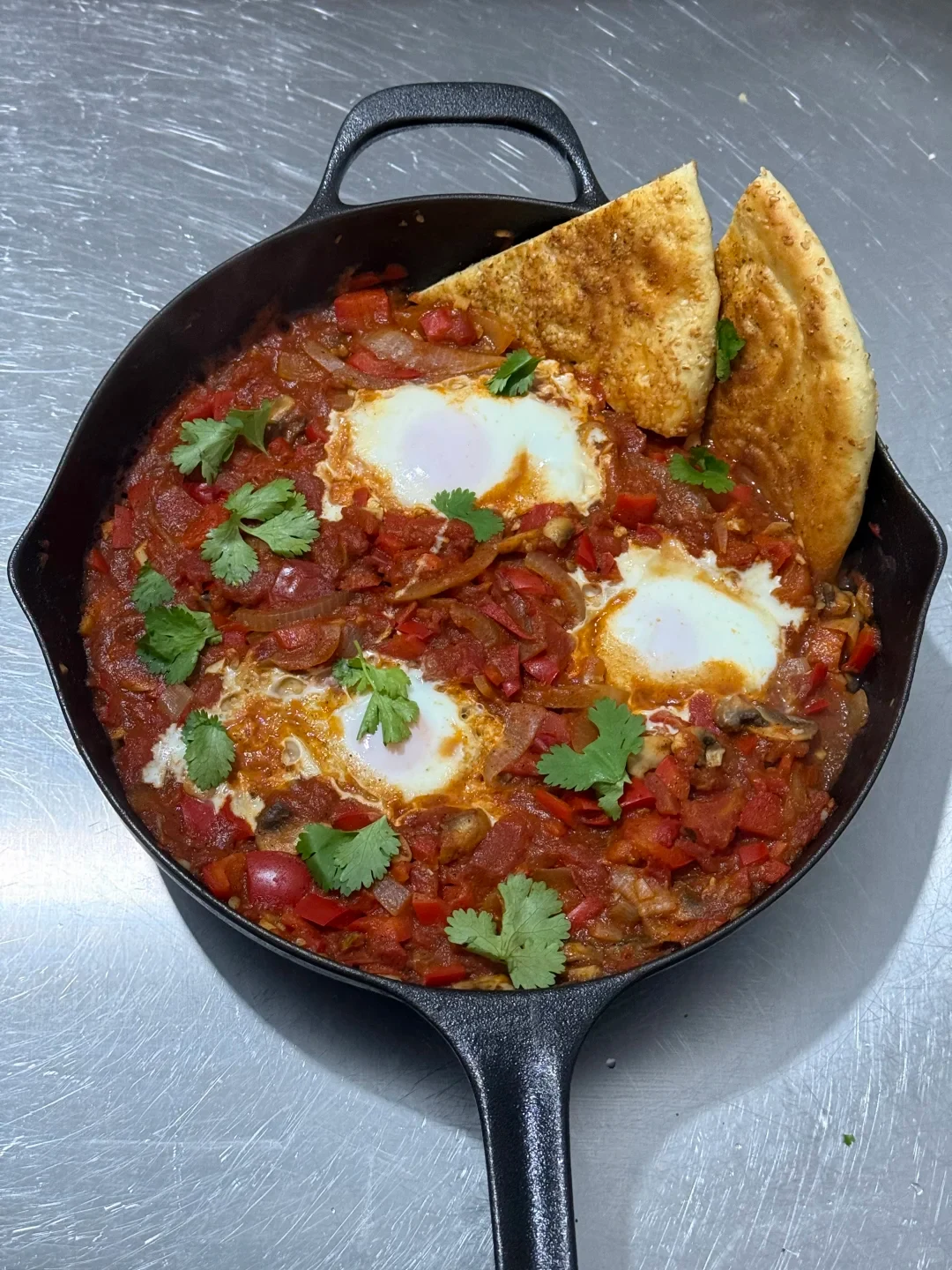 A black cast iron skillet filled with shakshuka, featuring poached eggs simmered in a rich tomato and bell pepper sauce, garnished with fresh cilantro leaves and served with toasted flatbread wedges on the side.