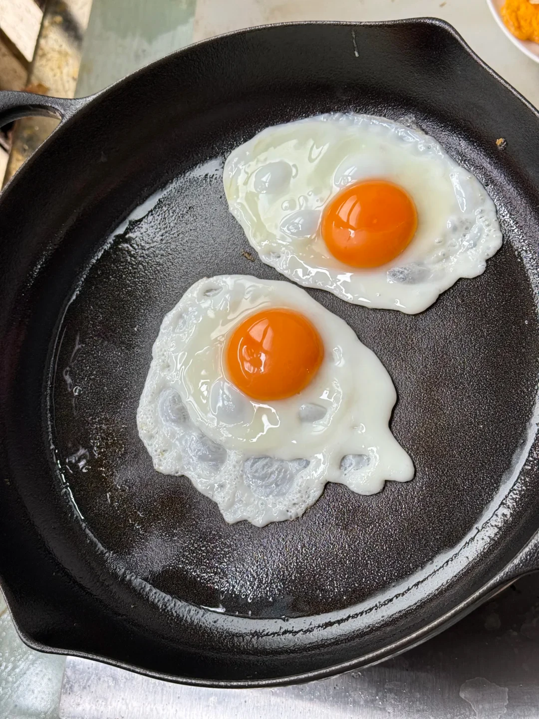 Two sunny-side-up eggs frying in a black cast-iron skillet with bright orange yolks.