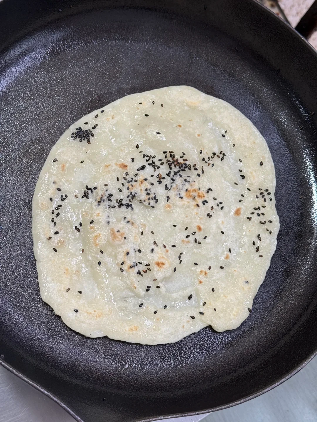 A thin flatbread sprinkled with black sesame seeds cooks inside a black cast iron skillet.