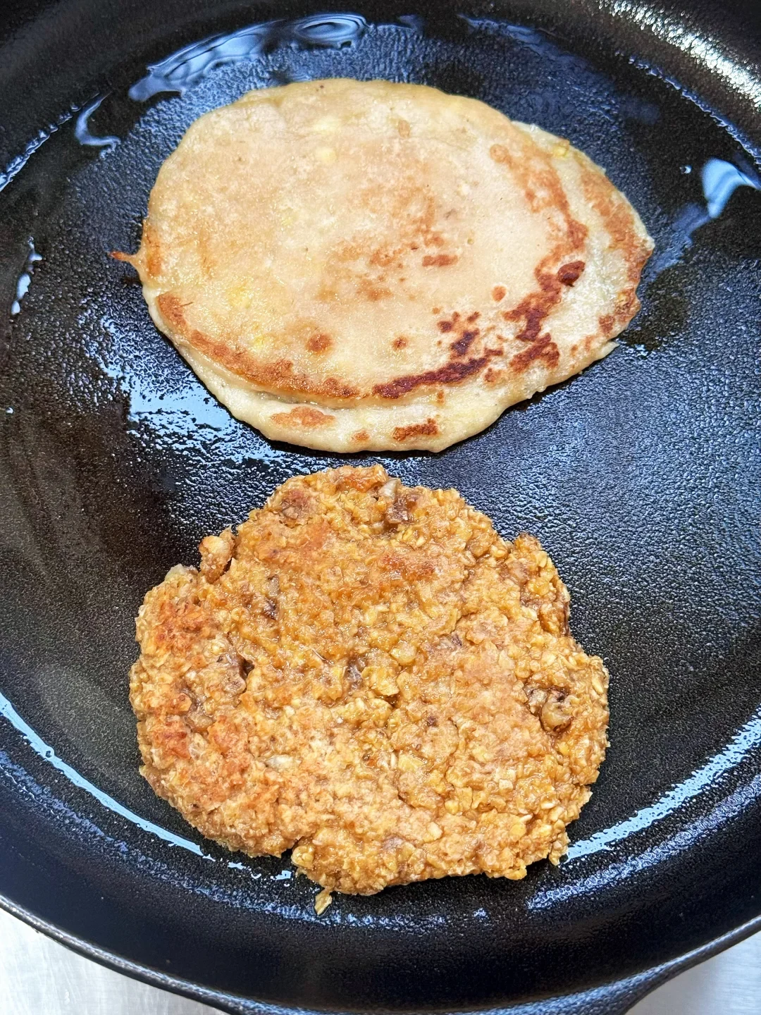 A smooth pancake and a coarse-textured patty frying in a black skillet.
