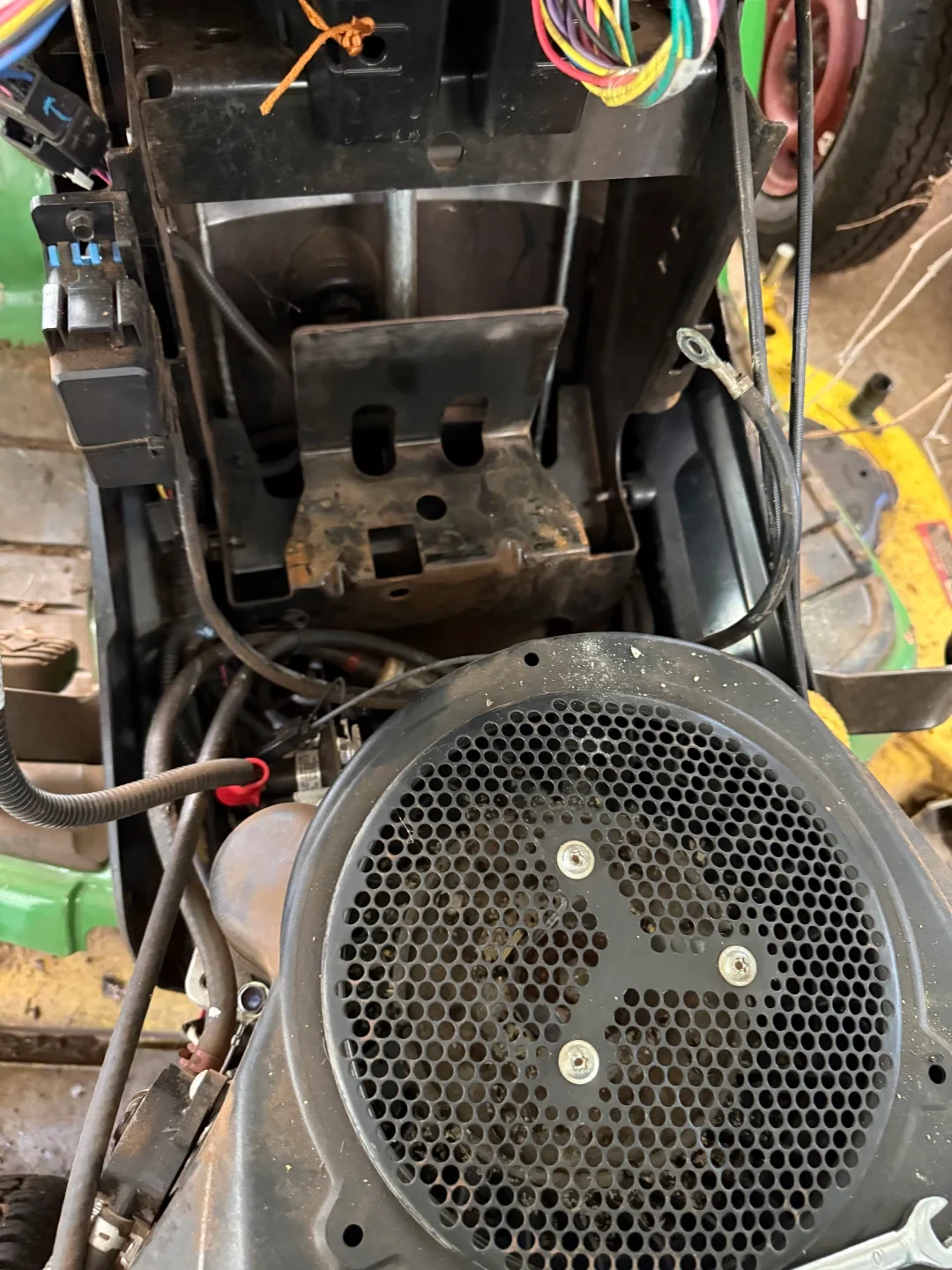 Top-down view inside the engine compartment of a riding lawn mower, showing an empty metal battery tray with disconnected cables next to the circular perforated engine shroud.