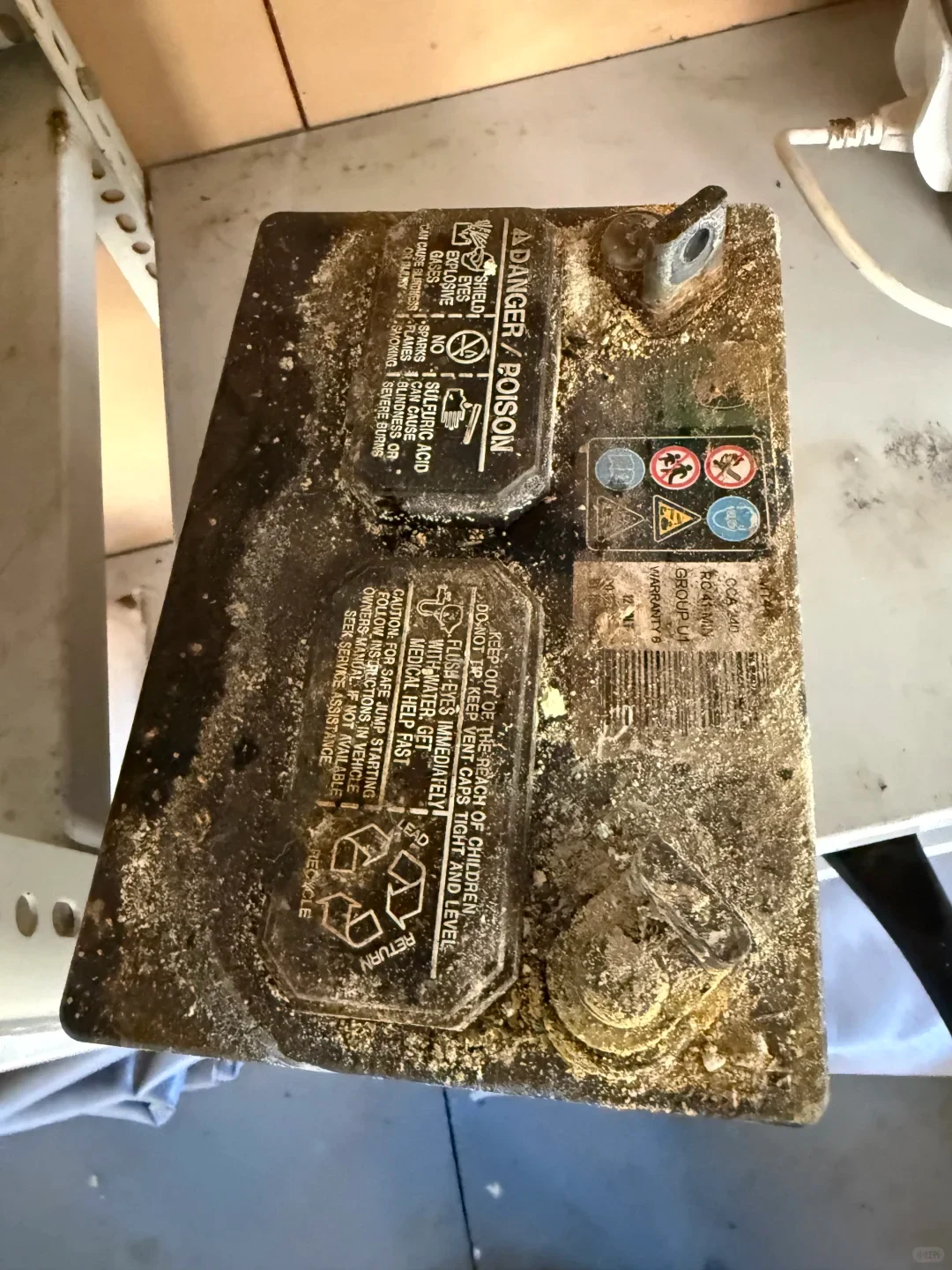 Close-up of a heavily corroded black lead-acid battery with white and yellowish residue, warning labels, and exposed terminals.