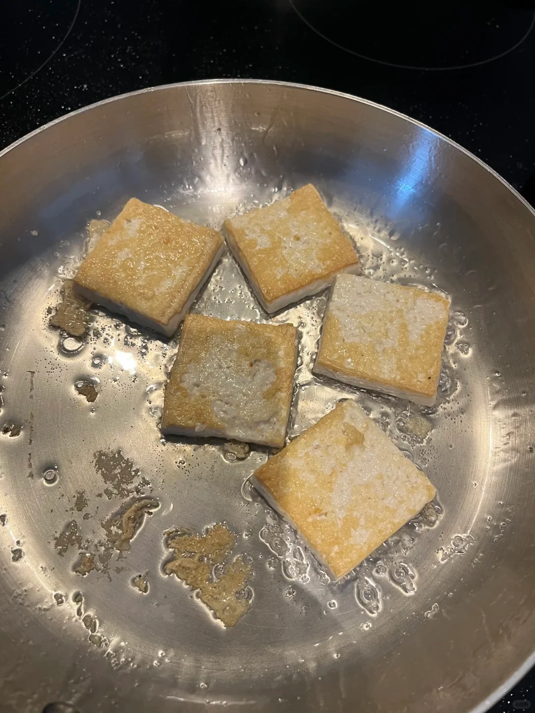 Five square pieces of tofu frying in oil inside a stainless steel skillet.