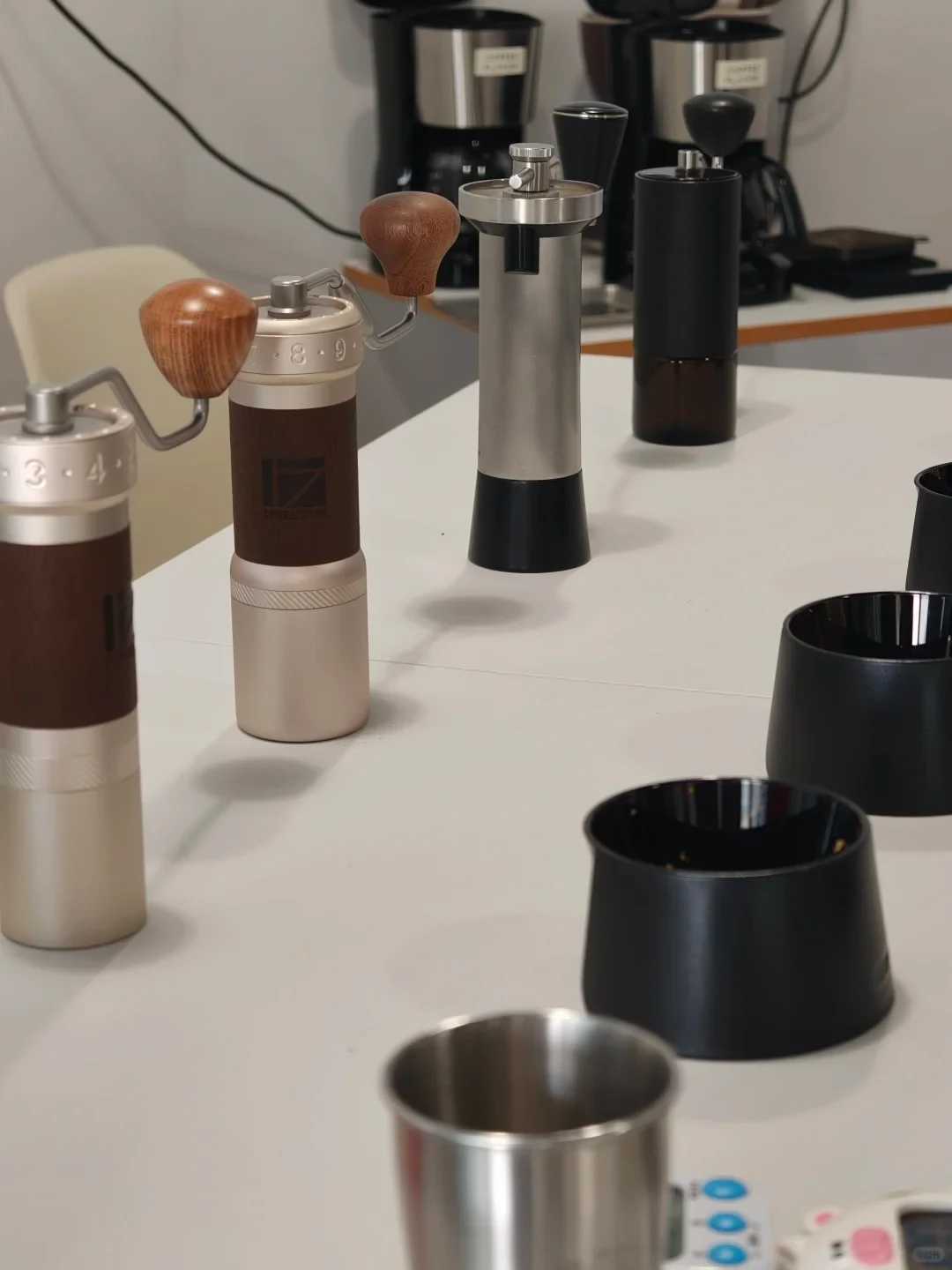 Several manual coffee grinders in different finishes, with wooden crank knobs and metal or dark cylindrical bodies, are displayed on a white counter alongside black and stainless-steel grinder cups. Drip coffee brewers sit in the background.