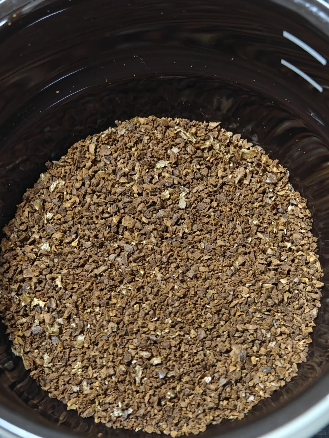 Close-up looking into a dark round container filled with coarse brown and tan flakes or granules.