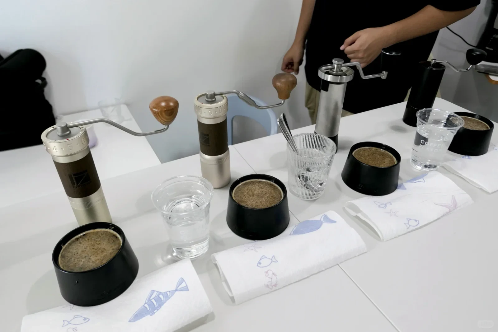 Several manual coffee grinders with wooden crank knobs are displayed on a white table beside cups of ground coffee, water glasses, and folded fish-patterned cloths; a person stands behind the table.