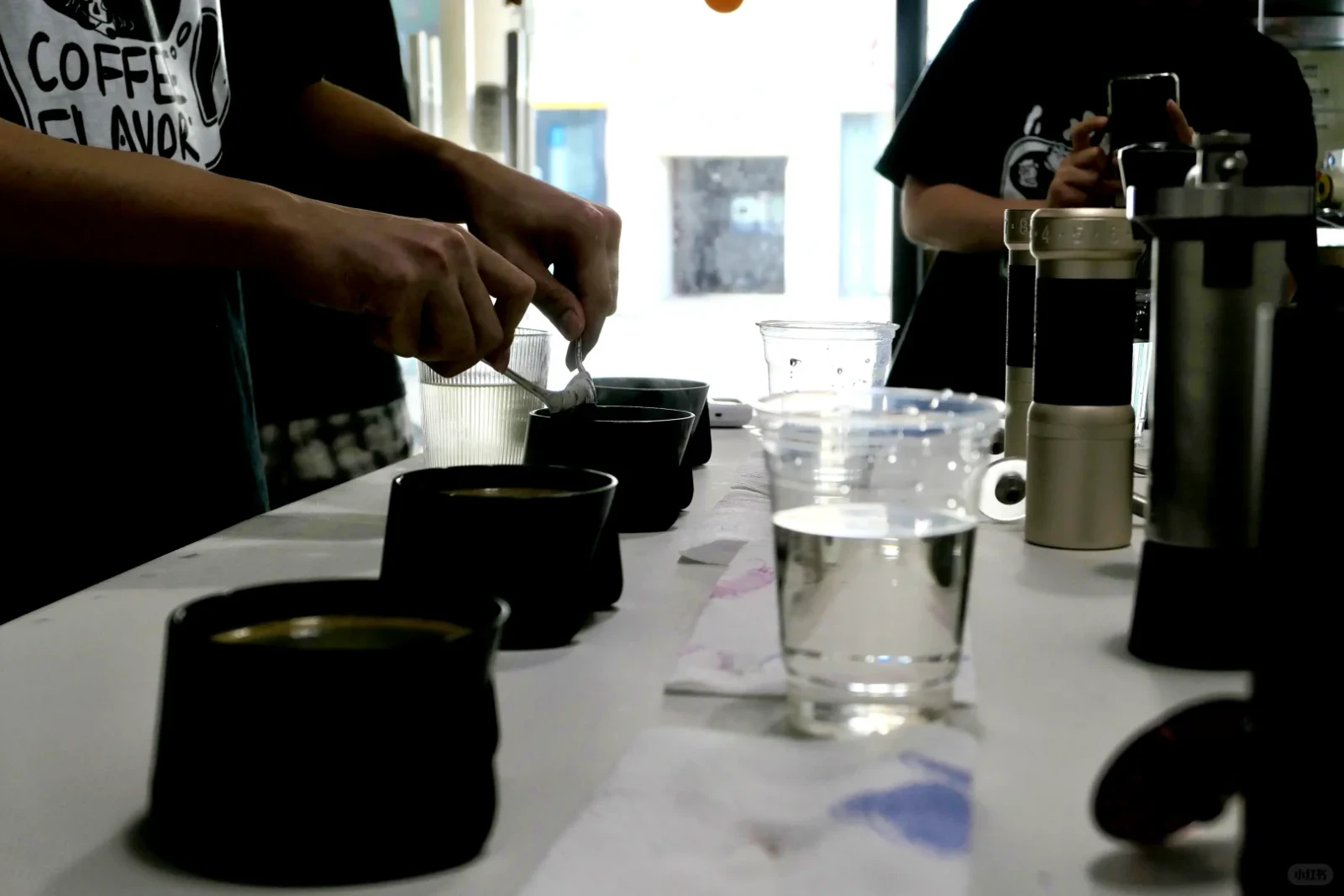 A person uses a spoon over a row of black cups at a coffee-tasting table, with water glasses and brewing equipment nearby; another person photographs the setup. A small watermark appears at the bottom right.