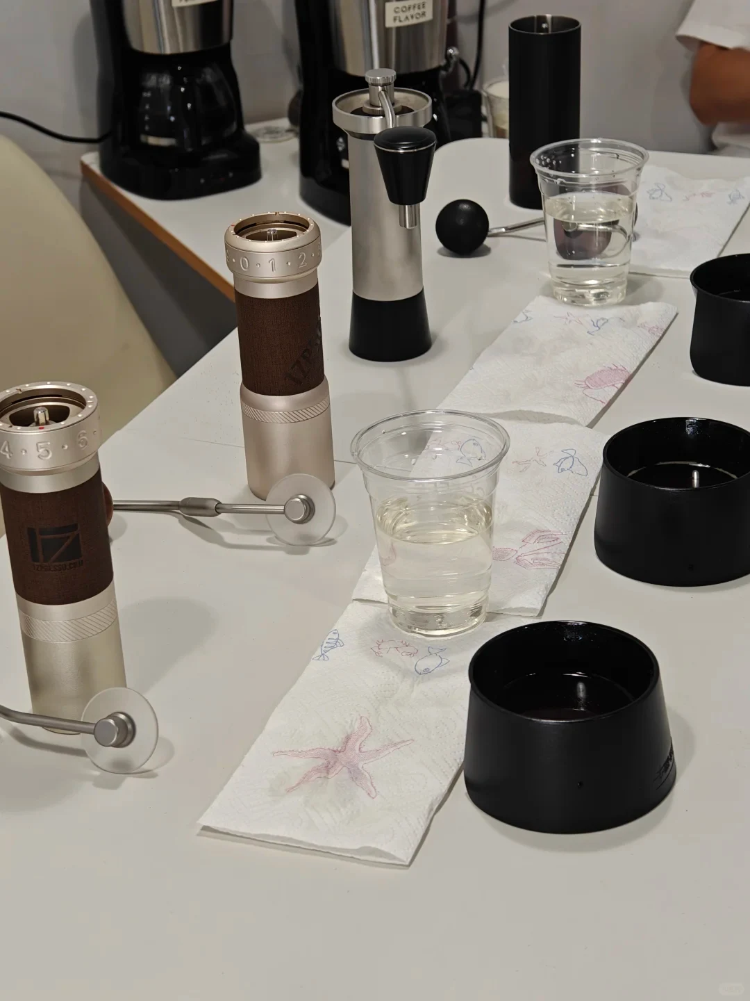 Several manual coffee grinders and disassembled grinder parts are arranged on a white table beside clear cups of water and patterned paper towels. Coffee machines sit in the background.
