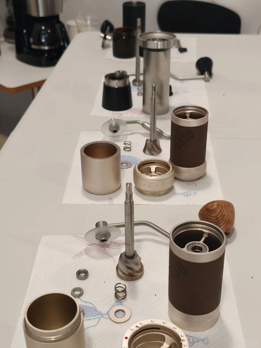 Disassembled manual coffee grinder components, including cylindrical housings, central shafts and burrs, springs, rings and catch cups, are laid out on paper towels on a table.