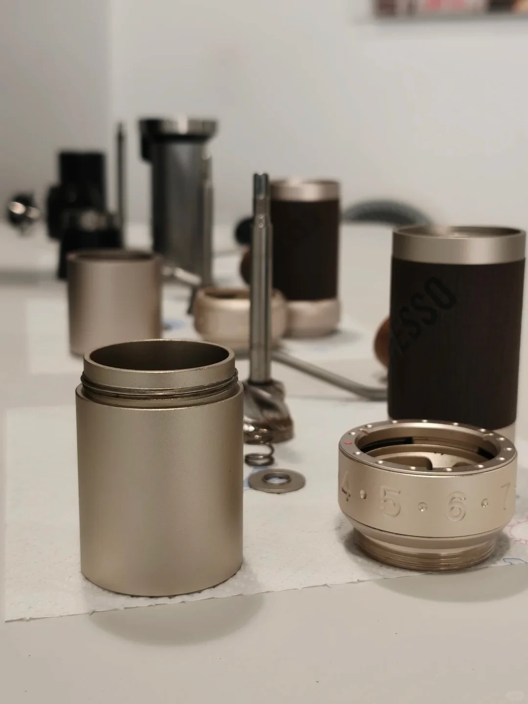 A disassembled cylindrical metal device and its parts are arranged on a tabletop, including an open body, a separate numbered adjustment ring, dark outer sleeves, and a central metal rod.