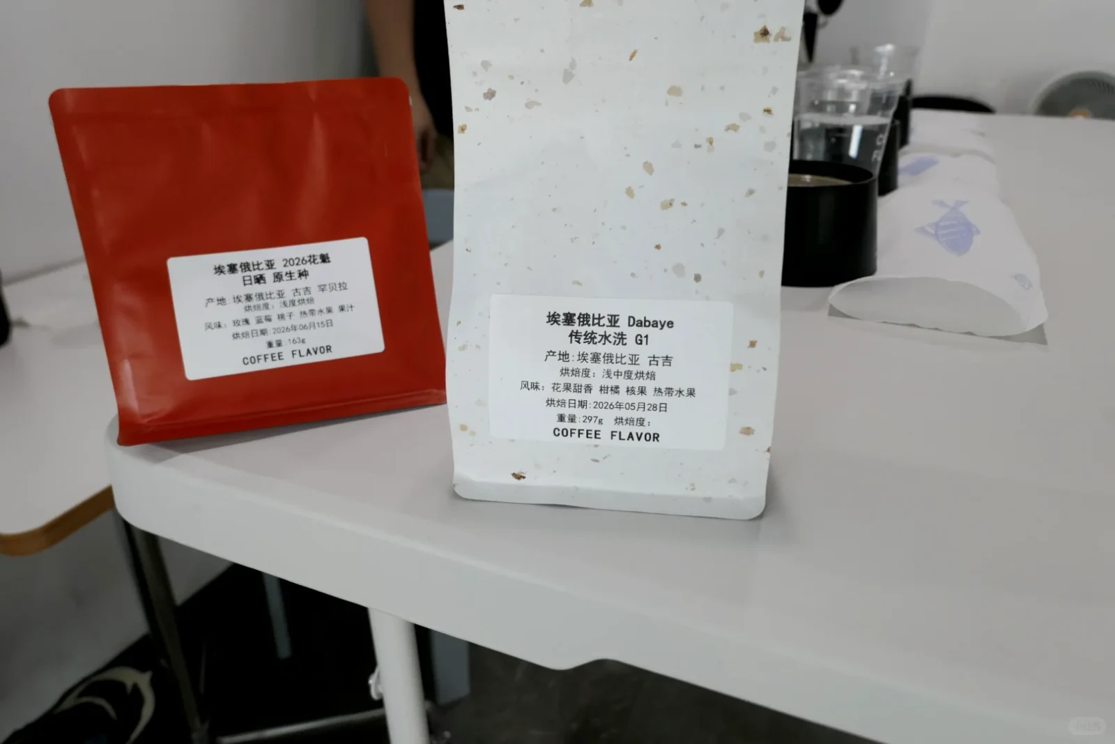 Two labeled coffee bags sit on a white table: a red bag on the left and a larger white speckled bag in front at center. Their labels identify Ethiopian coffee and list processing, origin, flavor notes, roast dates, and weights. A coffee brewer and paper filter are visible behind them.