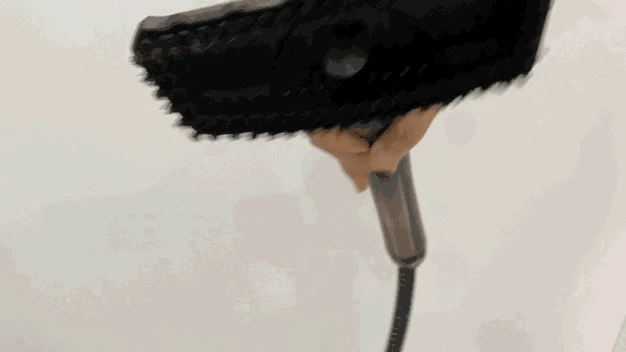 A person wiping a white tiled wall with the handheld bristle brush attachment of a steam cleaner.