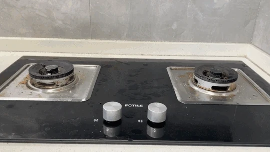 Dirty two-burner black glass gas stovetop with grease stains around the metal burner trays before cleaning.