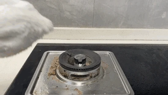 Handheld steam nozzle covered with a white microfiber cloth approaching a soiled stainless steel stovetop burner ring.