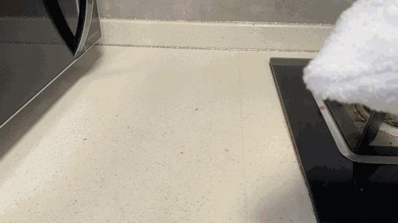 A handheld steam cleaner head covered in a white microfiber cloth being moved across a kitchen countertop between a microwave and a gas stove.