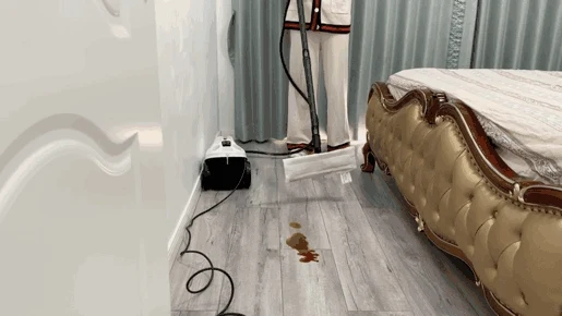 A person holding the floor nozzle of the Kärcher steam cleaner above a puddle of spilled sauce on a light wood floor next to an ornate bed.