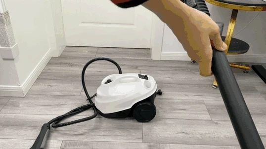 A hand holding the extension wand of a Kärcher SC3 steam cleaner with the wheeled base unit resting on a gray wood-look floor.