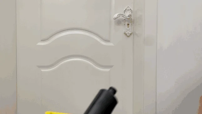 A black steam nozzle attached to a yellow steam cleaner body pointed toward a white interior door with an ornate handle.