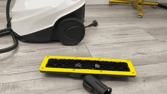 Floor nozzle attachment of the Kärcher SC3 steam cleaner lying upside down on a wood-look floor, with the main wheeled steam cleaner unit in the background.